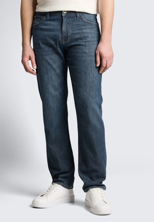 Personne portant un jean bleu droit, des baskets blanches et un haut beige, debout devant un fond clair uni.