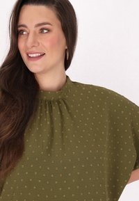 Oliwkowa bluzka z wysokim kołnierzem, krótkimi bufkami i teksturowanym wzorem małych złotych kropek. Miękka tkanina z marszczeniem przy dekolcie.