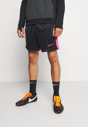 Mand iført sorte og pink Nike sportsshorts, sorte sokker og sorte Nike fodboldstøvler med klare orange snørebånd, stående mod en hvid baggrund.