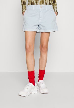 Person trägt hellblaue Shorts, rote Socken, weiße Sneakers und ein grünes gemustertes Oberteil und steht mit den Händen in den Taschen.