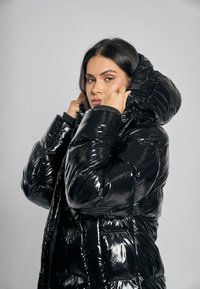 Czarna puchowa kurtka o błyszczącym wykończeniu, z dużym kapturem, pikowanym wzorem i elastycznymi mankietami; gładka tekstura i ciepła izolacja.