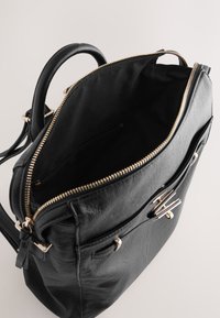 Zwarte leren tas met een ritssluiting aan de bovenkant, gouden hardware en een binnenzak met rits. Heeft een gestructureerde vorm en dubbele handvatten.