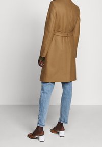 Manteau couleur chameau avec une coupe cintrée, une taille ceinturée et des manches longues. Associé à un jean bleu clair et des mules marron à talons.