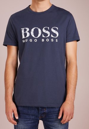 Mężczyzna w granatowym krótkim rękawie T-shircie z dużym białym napisem "BOSS HUGO BOSS", w połączeniu z niebieskimi dżinsami, na jednolitym tle.