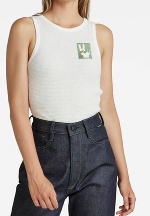 Kobieta w białym, bezrękawnikowym, teksturowanym topie z zielonym logo dłoni na piersi oraz w ciemnych dżinsach z wysokim stanem, stojąca z rozluźnionymi ramionami.