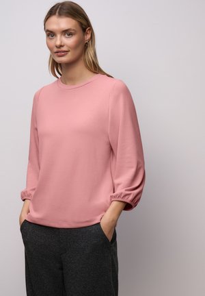 Haut rose pâle à manches longues avec un col rond et des manches bouffantes. Le matériau semble doux, assorti à un pantalon foncé texturé.