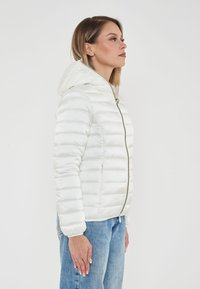 Giacca puffa bianca con design trapuntato, dotata di cappuccio, zip anteriore e polsini elasticizzati. Indossata su jeans azzurri chiari.