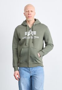 Olivovo zelená mikina na zips s prednými vreckami a kapucňou so šnúrkou. Obsahuje biele logo na hrudi. Nosené s svetlomodrými džínami.