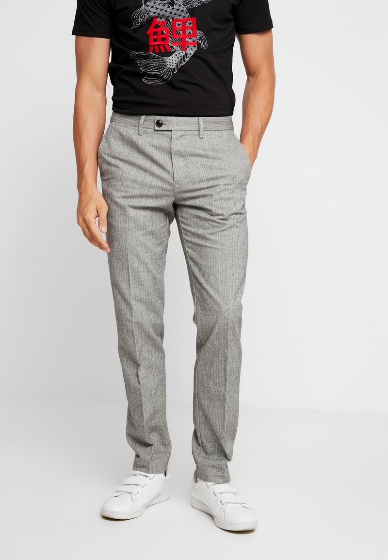 Homme portant un pantalon gris clair slim, un t-shirt noir avec un motif de poissons koï rouges et blancs, et des baskets blanches, debout avec une main dans la poche.