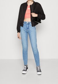 Chaqueta bomber negra sobre un top corto color coral con un corte en forma de corazón, combinado con jeans de tiro alto azul claro y zapatillas negras altas.