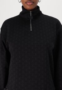 Pull over noir à col haut avec zip et motif répétitif "HUGO". Le tissu a une texture lisse et une coupe décontractée.