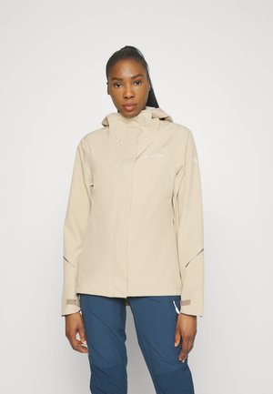 Giacca impermeabile beige con cappuccio, dotata di una zip frontale, polsini regolabili e dettagli riflettenti, abbinata a pantaloni blu navy.