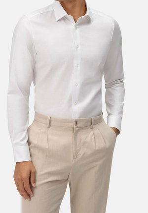 Mężczyzna ubrany w dopasowaną białą koszulę zapinaną na guziki, wsuniętą w beżowe spodnie z zakładkami, z jedną ręką w kieszeni, a drugą luźno opadającą.