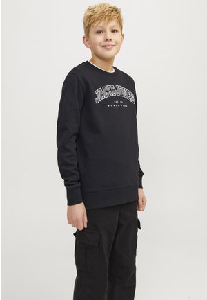 Blond chlapec v čiernej mikine s nápisom "JACK & JONES" a čiernych cargo nohaviciach stojaci pred jednoduchým svetlým pozadím.