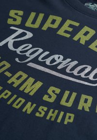 Camiseta de tela en color navy con texto en negrita y multicolor: "SUPERB", "Regional" y "Pro-Am Surf Championship" en amarillo y plata.