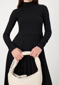Mujer vistiendo un vestido negro de manga larga sosteniendo un bolso crema con un asa acolchada y la marca "TOMMY" en la correa.