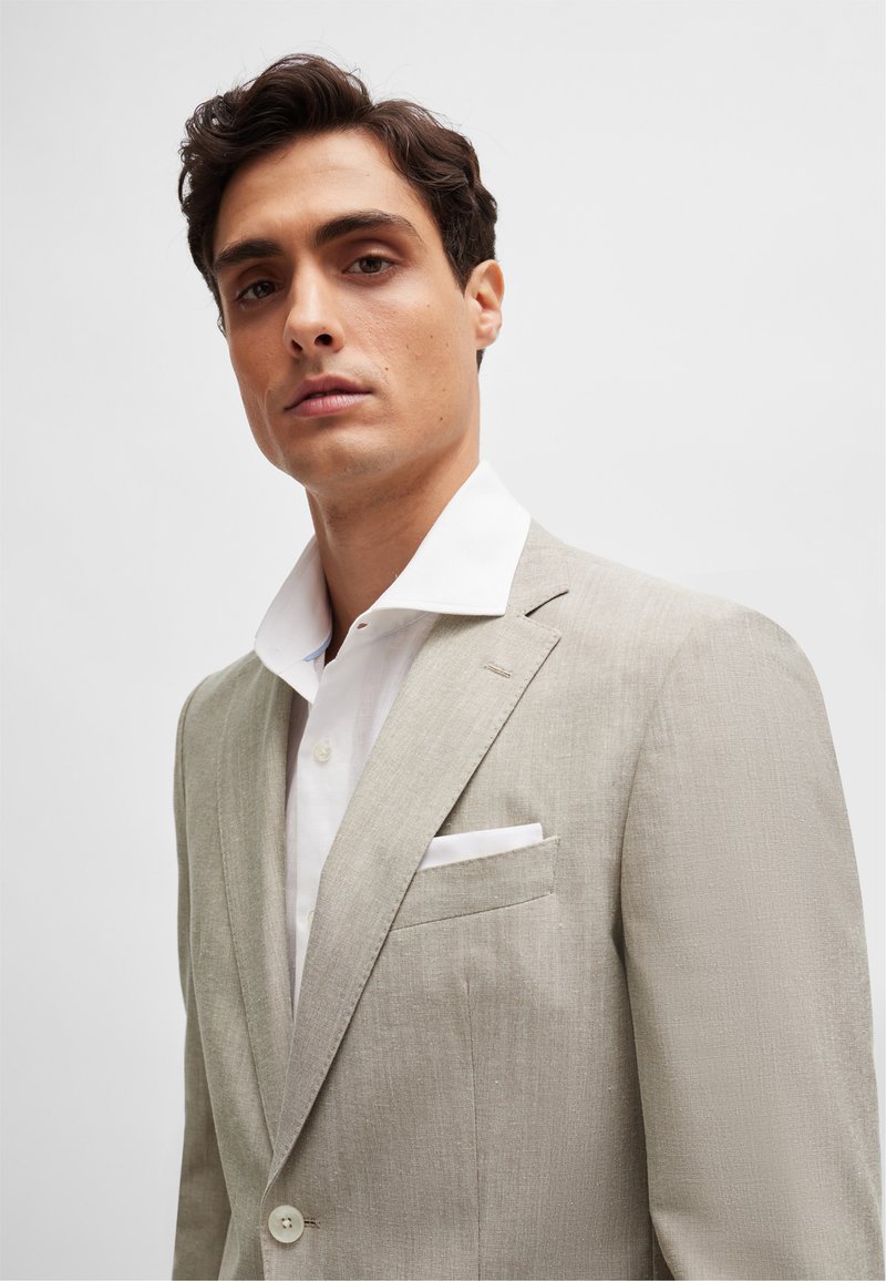 Blazer in lino beige chiaro con revers, camicia bianca sotto e un fazzoletto da taschino nella tasca sul petto, texture liscia.