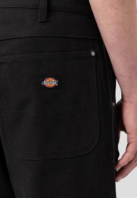Svarta canvastbyxor med en ficka, metallnitar och en röd och gul Dickies-logotyp. Har en kraftig textur och midjeband.