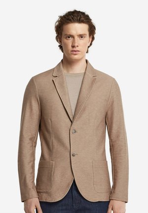 Beige Blazer aus weichem Material, mit aufgeschlagenem Revers, zwei Fronttaschen und Zwei-Knopf-Verschluss. Getragen über einem hellen Hemd.