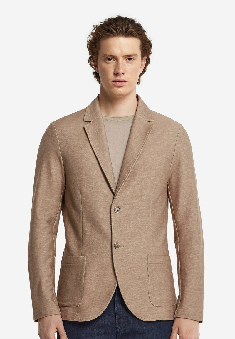 Beige kavaj med mjuk textur, med snedskuren krage, två framfickor och tvåknapps stängning. Bärs över en ljus skjorta.