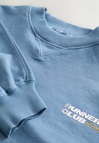 Felpa azzurra chiara con polsini e scollatura a coste, con il logo "RUNNER CLUB" in bianco e giallo. Texture morbida, design casual.