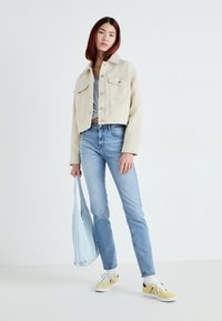 Casaco creme curto com bolsos frontais, blusa cinza riscada, jeans azul claro, sapatilhas amarelas e uma bolsa de mão azul pálido.