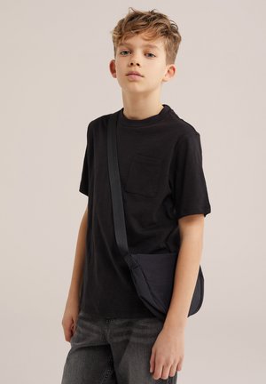 Jauns puisēns ar cirtainiem matiem valkā melnu t-kreklu un pelēkas džinsa bikses, nēsā mazu melnu somiņu pāri plecam uz vienmērīga bēša fona.