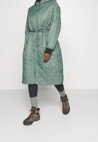 Persona che indossa un lungo cappotto trapuntato verde con cintura in vita, leggings neri, calze grigie e scarponi da trekking marroni, in piedi contro uno sfondo semplice.