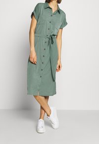 Donna che indossa un vestito midi verde con bottoni, maniche arrotolate e sneakers bianche, in piedi con le gambe incrociate su uno sfondo semplice.