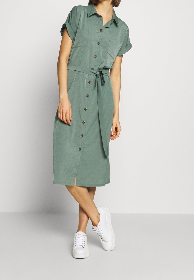 Donna che indossa un vestito midi verde con bottoni, maniche arrotolate e sneakers bianche, in piedi con le gambe incrociate su uno sfondo semplice.