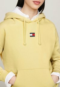 Felpa gialla in cotone con tasca frontale, cordino regolabile e una piccola patch con il logo "TOMMY JEANS". Indossata sopra una maglietta bianca.