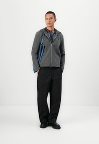 Grå hettegenser med glidelås og frontlomme, lagd over en mørk polo, sammen med svarte cargo-bukser og svarte sko, holder en denim totebag.
