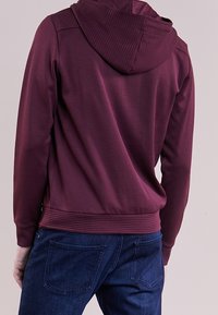 Sudadera burdeos con detalles acanalados en la capucha y el dobladillo, combinada con vaqueros azules con bolsillos traseros. Tejido suave, diseño casual.