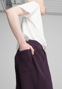 Balta trumpa rankovė t-shirt, derinama su tamsiai violetinėmis sportinėmis kelnėmis, kurios turi didelį šoninį kišenę ir elastingą juosmenį, fone naudojama neutralus spalvų tonas.