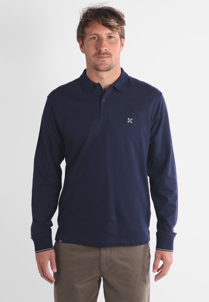 Homme portant un polo à manches longues bleu marine avec un petit logo brodé sur la poitrine, et un pantalon marron, se tenant devant un fond neutre.
