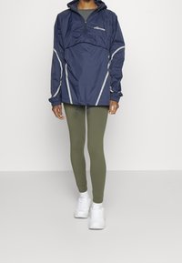 Mörkblå vindjacka med grå detaljer, halvdragkedja framtill, justerbar fåll, bärs över olivgröna leggings och vita sneakers.