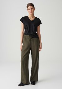 Schwarzes T-Shirt mit kurzem Arm und V-Ausschnitt kombiniert mit lockeren, olivgrünen, strukturierten Hosen mit elastischem Bund und Kordelzug. Schwarze Schuhe.