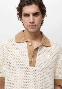 Cămașa polo tricotată are un corp crem, cu un guler și manșete texturate bej, închidere cu nasturi și un model triangular pe întreaga suprafață.
