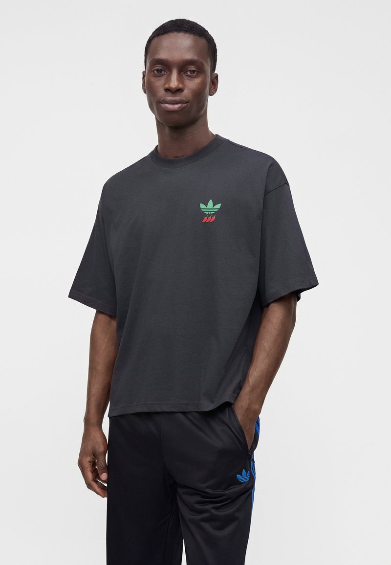 Young man wearing a loose black t-shirt with a small green and red logo and black pants with blue stripes and a blue logo.