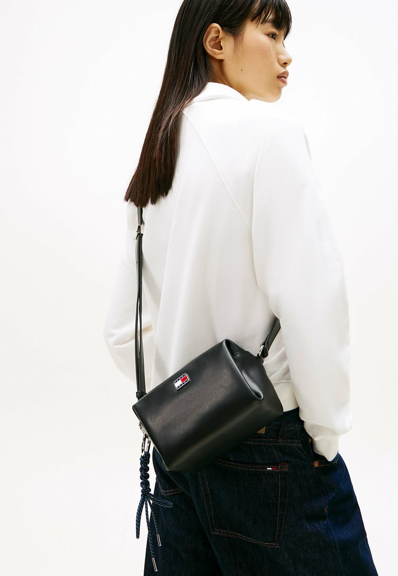 Ung kvinde med langt, mørkt hår iført en hvid sweatshirt og mørke jeans, bærende en lille sort læder crossbody-taske med logo-patch.