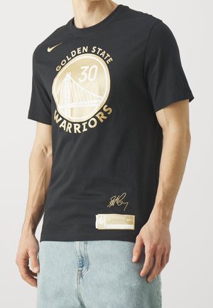 Homme portant un t-shirt noir des Golden State Warriors avec un logo doré, le numéro 30, une signature et un jean bleu clair, debout devant un fond blanc.