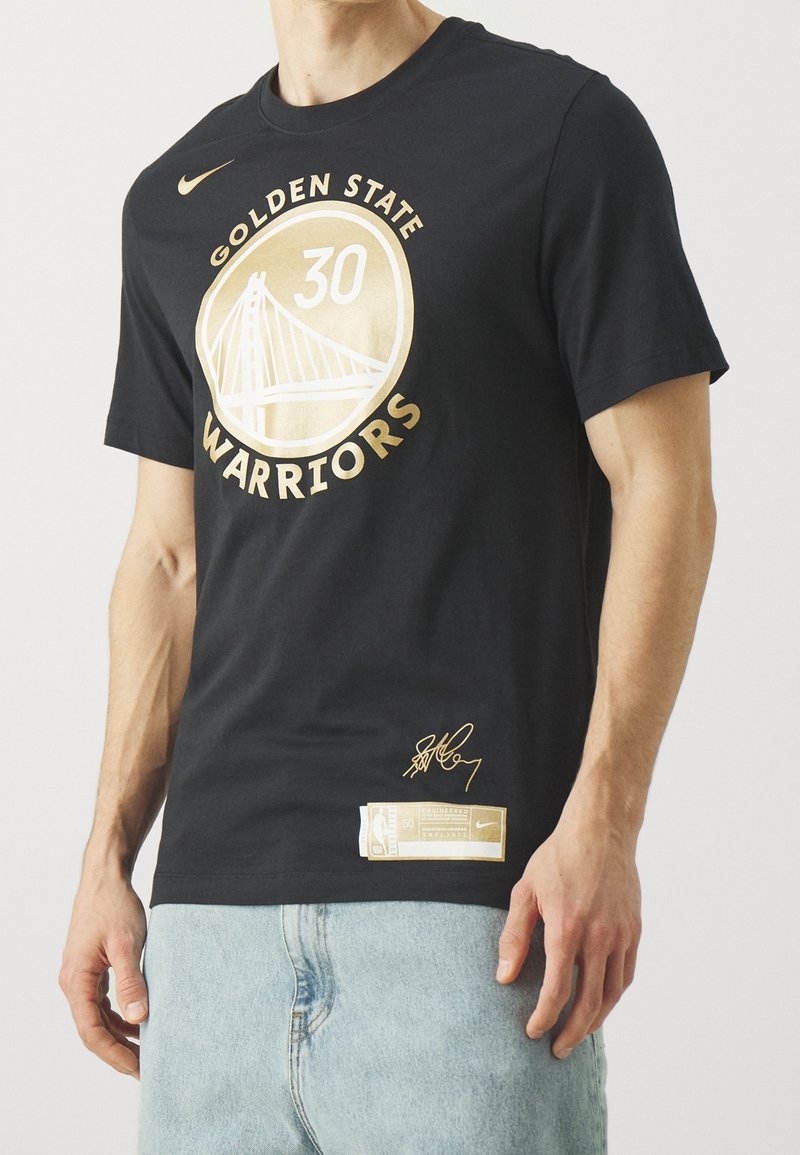 Homme portant un t-shirt noir des Golden State Warriors avec un logo doré, le numéro 30, une signature et un jean bleu clair, debout devant un fond blanc.