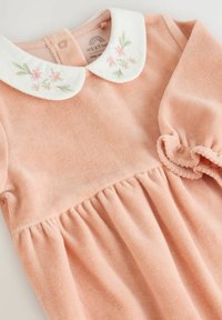 Robe en tissu rose doux avec un col blanc ornée de broderies florales. La taille froncée et les manches ballons ajoutent de la texture et du détail.