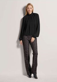 Schwarze Bluse mit hohem Kragen und voluminösen Ärmeln, kombiniert mit strukturierten, dunkel gemusterten Hosen und schwarzen spitz zulaufenden Schuhen.