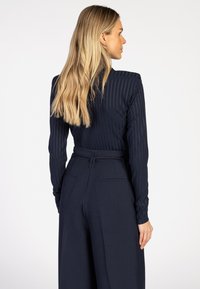 Vrouw met lang blond haar in een aansluitend, marine gestreept longsleeve-top en hoogtaille, wijde marine broek, kijkend naar rechts.