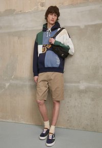 Sweat à capuche multicolore avec cordon de serrage, shorts beige et baskets. Présente des écussons en marine, vert, blanc et bleu avec du texte imprimé.