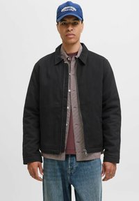 Giacca nera con zip frontale e colletto indossata sopra una camicia a quadri e una maglietta bordeaux; abbinata a jeans azzurri chiari.