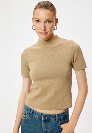 Lichtbruine top met korte mouwen en hoge hals. Textuurstof, aansluitende silhoeet. Gecombineerd met blauwe denim jeans.