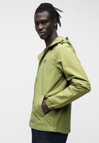 Νέος άντρας με ντραντλοκς που φοράει ανοιχτό πράσινο μπουφάν με κουκούλα και το χέρι στην τσέπη, στέκεται μπροστά σε απλό ανοιχτό φόντο.