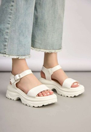 Pieds portant des sandales à plateforme épaisses blanches avec des brides à la cheville, associées à un jean bleu clair à ourlet effiloché sur un sol gris.